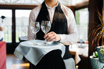 A young waiter in a stylish uniform stands with glasses on a tray near the table in a beautiful...