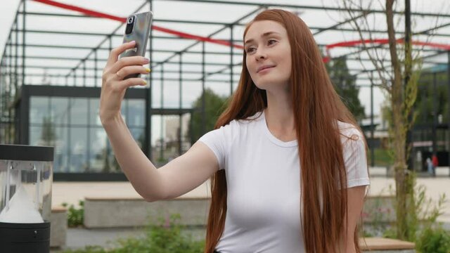 Beautiful ginger woman makes selfie on smartphone. Red hair attractive woman makes selfie photos on smartphone. Young lady poses in front of camera while sits on bench in park.