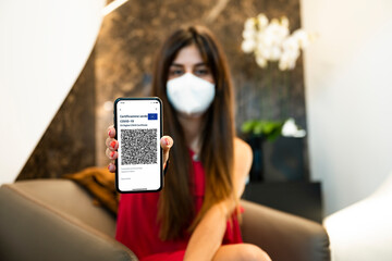 (Selective focus, focus on the smartphone) Defocused young girl, wearing a face mask, holding a passport and a smartphone with the Green Pass. 