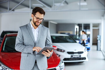 Smiling, friendly car seller standing in car salon and using tablet to check on new messages...