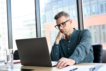 Focused middle aged businessman corporate leader ceo executive manager thinking of online planning, business analyst analyzing financial market strategy using laptop computer sitting at office table.