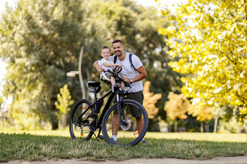 Fototapeta premium Father and son ride bicycles. The toddler is sitting in the basket of the bicycle while his dad is holding him while he is resting from riding the bicycle through the forest. Summer sunny family day