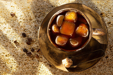 Cubes of frozen coffee in a ceramic cup, next to a cane sugar cube in tongs on a saucer and coffee beans on a textured background.  Coffe convenience. Top view, copying space.