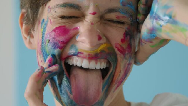 Young woman with rainbow colored face and hands smiles and shows her tongue. Portrait of happy gay girl posing emotionally and looking at camera