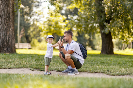 The family is having fun in the park. Father and son are playing in the forest dressed in the same casual summer clothes on a beautiful sunny day. Dad squats as the boy hi five to him. Making memories