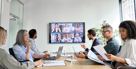 Fototapeta Diverse company employees having online business conference video call on tv screen monitor in board meeting room. Videoconference presentation, global virtual group corporate training concept. obraz