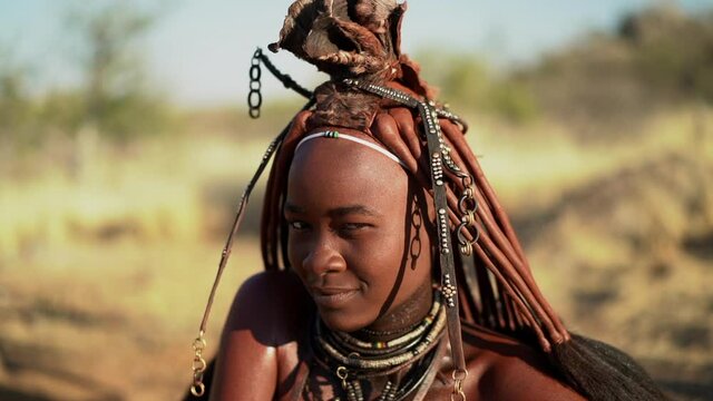 Beautiful Himba woman smiling, wearing traditional jewellery and headdress in her village near Kamanjab in northern Namibia, Africa, slow motion shot. 
