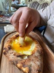 A man's hand dips a piece of khachapuri into the yolk on top of the dish. 