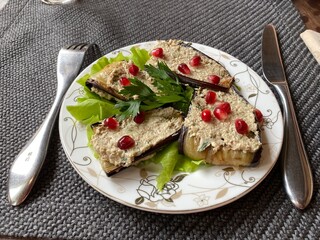 A sprig of parsley is decorating a dish of Georgian cuisine made of eggplant, nut paste and pomegranate seeds. The plate stands on a woven gray tablecloth. Next to the plate are a fork and a knife. 