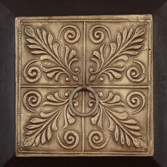 a square, decorative stamped metal panel of a dark wooden drawer with stylized botanical motifs and a circular pull in the center, in brown tones
