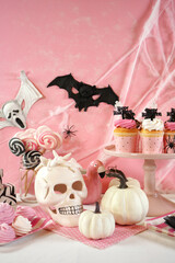 On trend pink Happy Halloween children's party table with cupcakes, white pumpkins, skull, spiders, and spooky decorations. Full table setting vertical orientation.