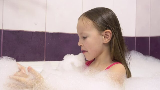 Little girl in a pink bathing suit with a lot of foam in the bath. She blows the foam off her hands.