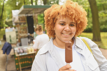 Lifestyle portrait of smiling young naturally beautiful plus size African American woman with afro red hair eating ice cream chocolate popsicle in public park. Diversity and body positive concept