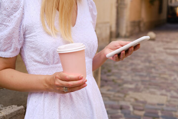 Girl drinks coffee from a pink paper cup and uses a smartphone. A woman is holding a smartphone, sending a text message, or using an app on her mobile phone. Drink cappuccino or latte in the street