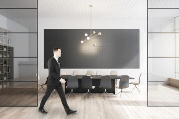 Businessman walking in modern meeting room interior with large table, wooden flooring, equipment, glass windows and sunlight.