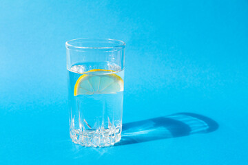 Fresh clean water in a glass with a slice of lemon on a blue background in bright sunlight. Live water, healthy drink