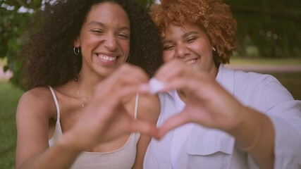 Young beautiful happy lesbian African American couple sitting on green grass showing love sign outside at nature summer park. LGBT community concept. Female friends smiling enjoying moments together.