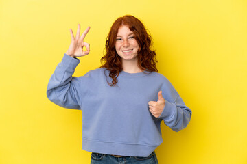 Teenager reddish woman isolated on yellow background showing ok sign and thumb up gesture