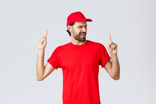 Upbeat Handsome Bearded Delivery Guy Spending Break Relaxing, Employee In Red Uniform Cap And T-shirt Singing Song, Pointing Fingers Up Carefree, Close Eyes, Standing Grey Background