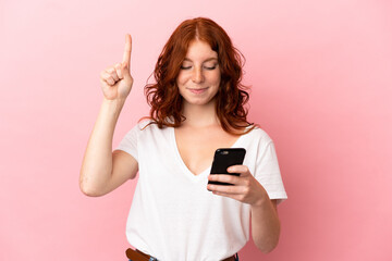 Fototapeta premium Teenager reddish woman isolated on pink background using mobile phone and lifting finger