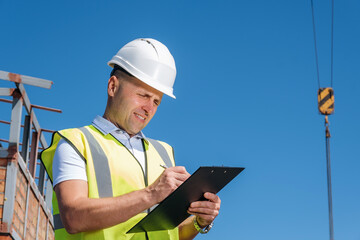 Mature man in white helmet stands on the roof, holds clipboard, inspects construction site and writing notes on blue sky background, copy space for text