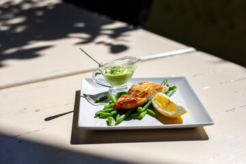 Fried fish with green beans, lemon and green sauce on white square plate on the table outside