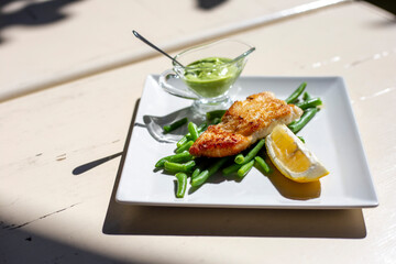 Grilled salmon with green beans, lemon and green sauce on white square plate on the table outside. Fried fish