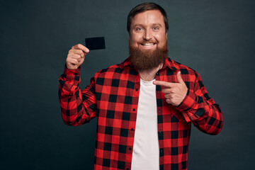 Portrait of a cheerful bearded man pointing finger at credit card isolated over grey background