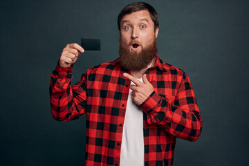 Portrait of a shocked bearded man pointing finger at credit card isolated over grey background