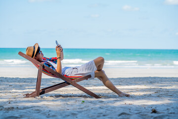 Young Asian man resting on sunbed on tropical beach. Happy guy sit on beach chair by the sea using...