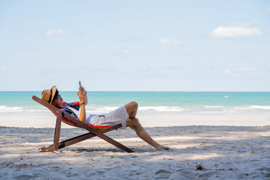 Young Asian man resting on sunbed on tropical beach. Happy guy sit on beach chair by the sea using smartphone for selfie or video call. Handsome male enjoy beach outdoor lifestyle on summer vacation
