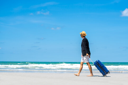 Happy Asian businessman tourist in casual suit barefoot walking on tropical beach with blue suitcase luggage in summer sunny day. Handsome man enjoy outdoor activity lifestyle on summer vacation