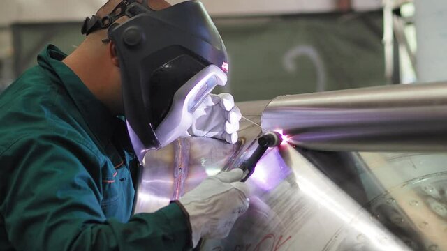 Skilled welder using TIG welding machine inside workshop, wearing mask. He is welding the stainless steel pipes in the factory. Side view static shot.