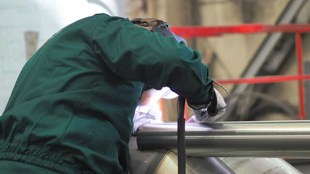 Skilled welder using TIG welding machine inside workshop, wearing mask. He is welding the stainless steel pipes in the factory. Shot from the back.
