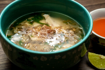Close-up photo of a bowl of Soto Seger, one of the Indonesian specialties, made from chicken in a soup-like sauce, accompanied by chili sauce, lime juice, and a wooden textured background