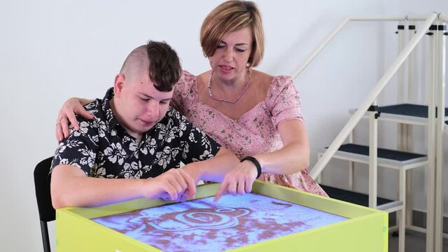 Children with disability getting sensory activity Sand tray, sand table for animation, cerebral palsy boy playing calming game, training fine motor skills. Rehabilitation center with therapist, mother