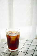 Cold brew cofee with ice on checked cloth beside a window with white curtain