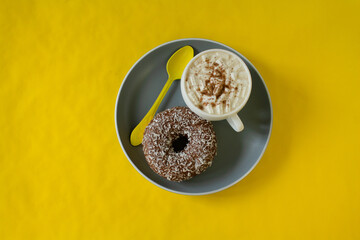 yellow background. a white cup, a gray plate with a donut, spoons.