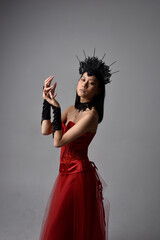 portrait of beautiful young asian woman wearing red corset and crown, posing in gothic horror style with creative hand gestures isolated against dark black studio background.