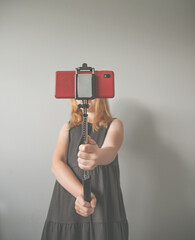 girl in a gray dress holds a selfie stick with a red phone covering her face on gray background. Concept without face