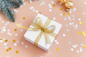 Obraz na płótnie Canvas christmas gift box golden color ribbon on beige background with confetti, new year decoration balls and fir tree branches