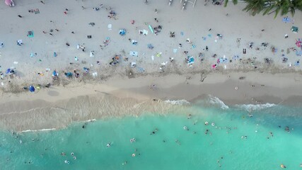 A drone hovers above a crowded tropical beach vacation destination as families enjoy sun, sand and surf together.