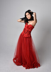 Full length  portrait of beautiful young asian woman wearing red corset and ornate gothic queen crown. Graceful standing posing  isolated on studio background.