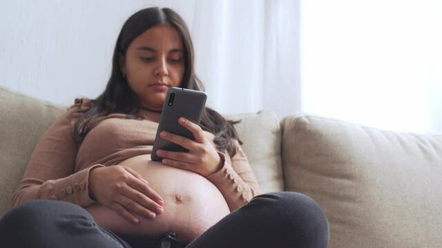 A young pregnant hispanic woman sitting on the couch with the smart phone