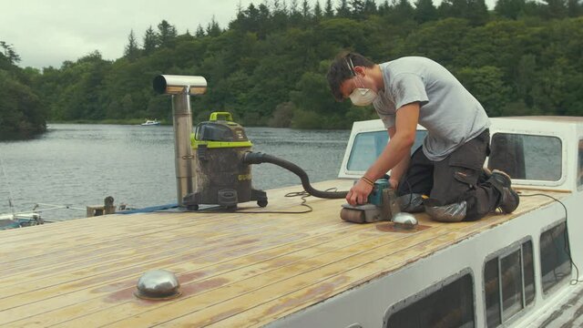 Young man belt sanding planks on wooden boat forecabin. WIDE SHOT
