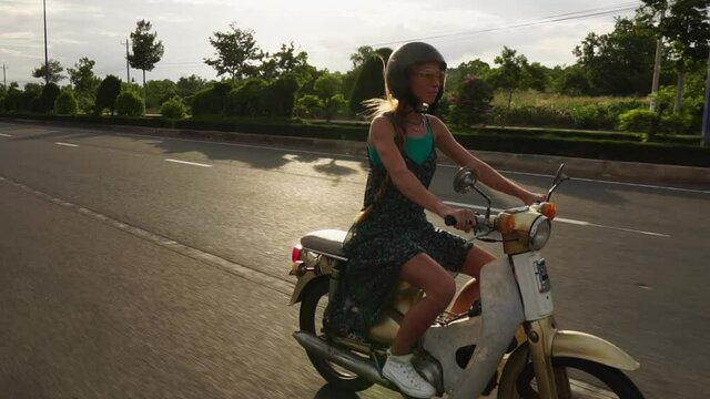 Young Caucasian woman with long blond hair cautiously riding a vintage style moped along the deserted roads of Vietnam.