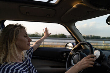 Fototapeta na wymiar Relaxed calm woman driving car enjoy speed and freedom stretching hand in open window. Confident middle age female driver look at horizon sunset and road. Blonde casual girl in 40s hold steering wheel