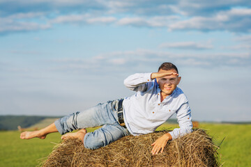 Naklejka na ściany i meble A young European lies on a bale of hay and looks ahead and smiles. Rest at nature. Portrait of a cheerful farmer in a shirt and jeans.