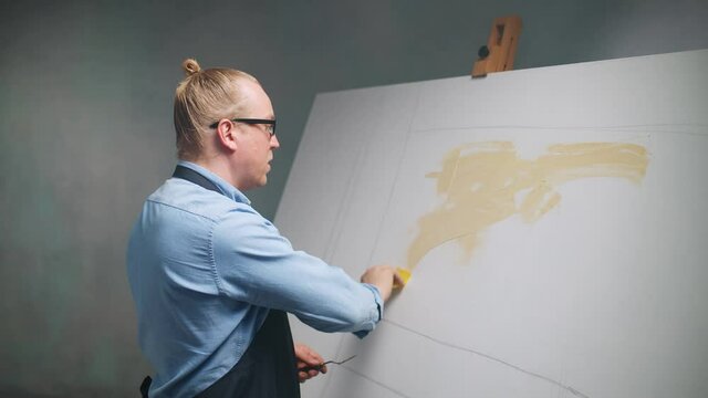 Slow motion, a talented adult man artist covers the canvas with beige oil paint, creative person at work, priming and draws the background.