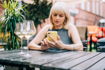 Adult mature woman texting messaging at mobile phone in bar outdoors with wine glasses and blurry restaurant, drinking white wine. People using technology cellphone. Lifestyle at summer sunny day 